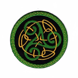 May include: A circular embroidered patch featuring a black background with a central green and gold triskelion knot design. The border consists of a green ring decorated with a repeating pattern of small, stylized gold leaf-like shapes.