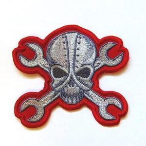 May include: Embroidered patch with a skull and crossed spanners design. The skull is detailed with metallic silver thread, and the spanners are silver. The patch has a red border, creating a striking contrast. Suitable for jackets, bags, or other fabric items.