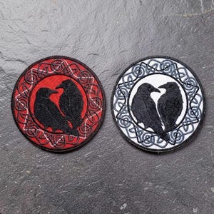 May include: Two circular patches featuring black raven silhouettes. One patch has a red background with a Celtic knot border, the other has a white background with a similar border. The ravens are facing each other.