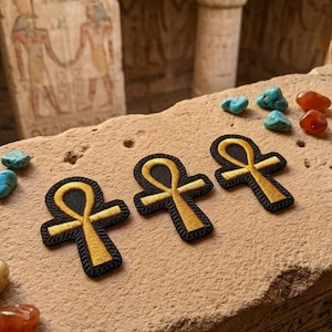 May include: Three embroidered Ankh patches with gold and black detailing, arranged on a textured surface. The patches are surrounded by small turquoise and orange stones, with an ancient Egyptian background.