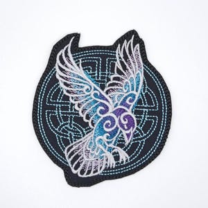May include: Embroidered patch featuring a stylized bird with outstretched wings. The bird is rendered in shades of blue, purple, and white against a black background with a Celtic knot design. The patch has a unique shape with a black border.