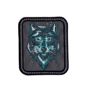 May include: A gray embroidered patch with a wolf's face in teal and white. The wolf's face is surrounded by a gray border with a decorative stitching pattern.