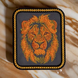 May include: A gray embroidered patch with a gold border featuring a lion's head with orange and yellow fur.