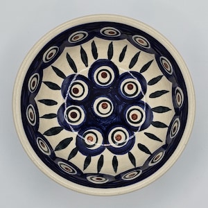 May include: A ceramic bowl with a cream exterior and a detailed interior design. The interior features a pattern of dark blue circles with brown centres, surrounded by green leaf-like shapes. The rim has a repeating pattern of blue and white circles.