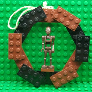 May include: A circular ornament constructed from alternating black and brown plastic building bricks, featuring a brown droid minifigure standing in the center. A light-colored string loop is attached to the top of the circular brick frame.