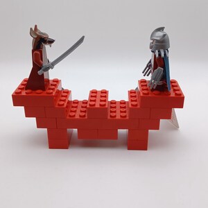 May include: Two plastic minifigures standing on a red brick structure. One figure is a brown rat-like character holding a gray sword, and the other is a silver-armored figure with a blue cape and clawed hands, facing each other on the platform.