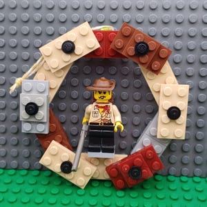 Puede incluir: Una minifigura de LEGO con sombrero de vaquero marrón, camisa beige con pañuelo rojo y pantalones negros, sosteniendo una varilla gris. La figura está colocada dentro de un marco circular construido con ladrillos LEGO y botones negros.