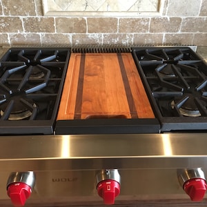 May include: A stainless steel gas stovetop with a wooden cutting board insert. The stovetop has four red knobs labeled "OFF" and the brand name "WOLF" is visible on the front.