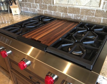 May include: A stainless steel gas stovetop with four burners and red knobs. A wooden cutting board is placed over two of the burners.