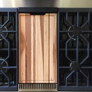 May include: A black and stainless steel stovetop with a wooden cutting board in the center. The stovetop has four burners and a black grate.