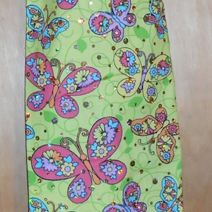 May include: A green reusable grocery bag with a pink drawstring and a floral pattern of butterflies with colorful accents. The bag is decorated with glitter.