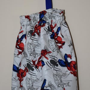 May include: A white drawstring bag with a blue strap. The bag is decorated with a repeating pattern of Spiderman characters in red, blue, and black.