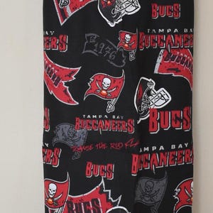 May include: A black and red reusable grocery bag with a repeating pattern of the Tampa Bay Buccaneers logo. The bag has a gray drawstring.