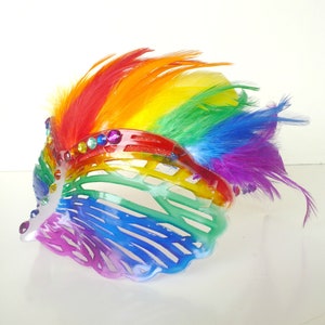 May include: A rainbow-coloured feather headdress with a clear plastic base and rhinestones. The headdress is made of feathers in the colours of the rainbow, with a clear plastic base that is decorated with rhinestones.