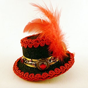 May include: A miniature black top hat with red trim and a matching feather. The hat features a gold band with red jewels and a large red jewel in the center. The hat is likely a decorative item or accessory.