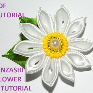 Puede incluir: Flor blanca Kanzashi con un centro amarillo y una perla. La flor está hecha de cinta de satén y tiene una hoja verde.