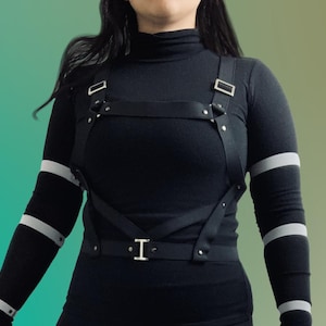 May include: A black elastic body harness featuring adjustable silver-toned metal buckles and rivets. The harness consists of multiple intersecting straps worn over a black long-sleeved turtleneck shirt with grey horizontal stripes on the sleeves.