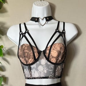 May include: A black strappy bustier featuring a beige and black floral print on the cups and sheer mesh bodice. The garment includes a matching black choker with a silver-toned metal heart-shaped ring at the center and adjustable shoulder straps.