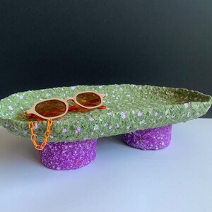 Op de afbeelding: Een groene en paarse papier-maché dienblad met een paar oranje zonnebrillen erop. Het dienblad heeft een gevlekt patroon en is gevormd als een skateboard.
