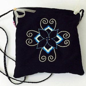 Könnte beinhalten: Eine marineblaue Umhängetasche mit floralem Design. Die Tasche hat eine zentrale gestickte Blume in Blau- und Weißtönen, umgeben von wirbelnden Mustern. Ein schwarzer geflochtener Riemen und eine weiße Schleife vervollständigen das Design.
