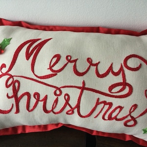 Könnte beinhalten: Ein rechteckiges, cremefarbenes Zierkissen mit roter, gestickter Schreibschrift, die Merry Christmas lautet. Das Kissen ist mit grünen Stoff-Stechpalmenblättern und roten Perlenbeeren in zwei Ecken verziert und hat einen roten Stoffrand.