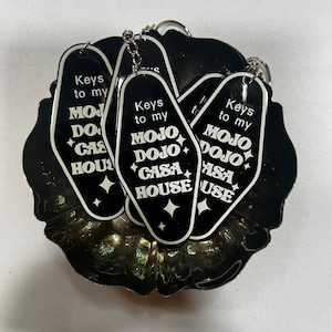 Puede incluir: Cinco llaveros negros con forma de llaves de habitación de motel con texto blanco que dice "Keys to my Mojo Dojo Casa House".