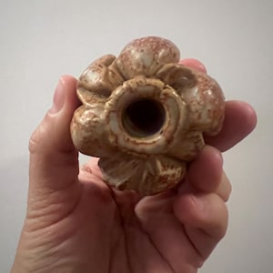 May include: A ceramic bottle stopper in the shape of a flower. The stopper is a mix of beige and brown with a textured surface. The stopper has a hole in the center.