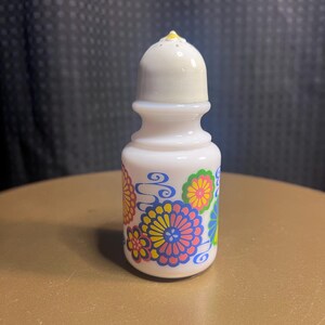 Op de afbeelding: Een witte keramische zoutstrooier met een bloemenontwerp in roze, geel, blauw en groen. De strooier heeft een afgeronde body en een gewelfde bovenkant met een klein geel accent. Het ontwerp doet denken aan vintage keukengerei.