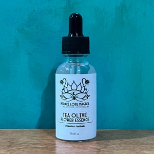 Può includere: Una bottiglia di vetro trasparente con contagocce nero, etichettata "TEA OLIVE FLOWER ESSENCE". L'etichetta presenta un disegno floreale e il testo "MERAKI LORE MAGICK & THE WONDER APOTHECARY". La bottiglia contiene un liquido trasparente ed è da 30 ml.