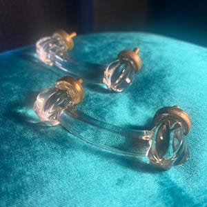 Könnte beinhalten: Vier transparente Glasschrankschubladengriffe mit messingfarbener Hardware. Die Griffe haben ein gebogenes Design mit einem dekorativen, strukturierten Ende. Die Griffe sind auf einer türkisblauen Oberfläche angeordnet.