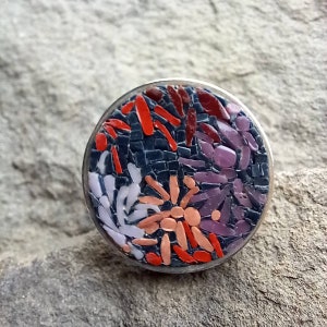 May include: A round, silver ring with a mosaic design. The mosaic is made of small, colorful tiles in shades of red, orange, purple, and white. The tiles are arranged in a floral pattern.