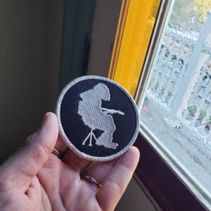 May include: A round, black embroidered patch with a white outline and a white silhouette of a person sitting in a chair. The patch is held up in front of a window with yellow and clear glass.