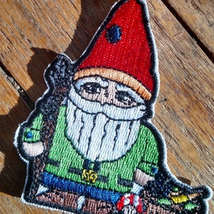 May include: Embroidered patch of a cartoon gnome wearing a red hat, green shirt, and blue pants. The gnome is holding a brown stick and sitting on a log.