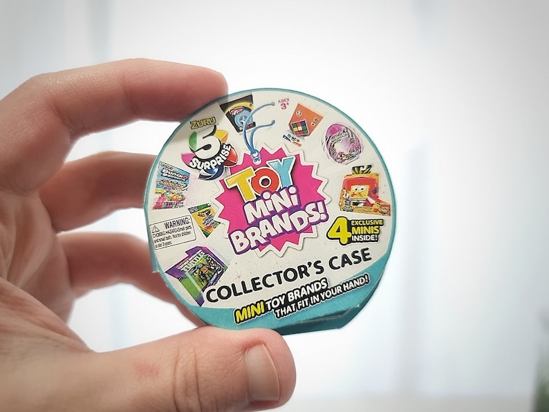 May include: A round, blue and white plastic container with the words "Toy Mini Brands! Collector's Case" printed on it. The container has a variety of small toy brands printed on it, including "Zuru 5 Surprise" and "4 Exclusive Minis Inside!"