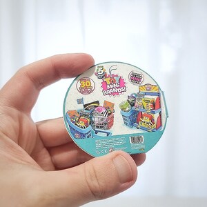 May include: A round, blue and white sticker with a picture of a miniature grocery store with a shopping cart full of miniature grocery items. The sticker says "Toy Mini Brands!" and "30+ Surprise Mini Toys!"