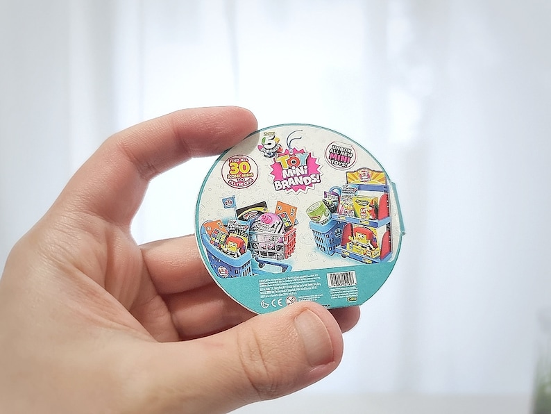 May include: A round, blue and white sticker with a picture of a miniature grocery store with a shopping cart full of miniature grocery items. The sticker says "Toy Mini Brands!" and "30+ Surprise Mini Toys!"