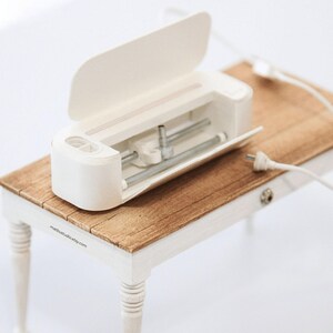 May include: A small, cream-coloured craft cutting machine with an open lid, displaying silver metal parts. It rests on a miniature wooden table with white legs. A white power cable is connected to the machine.