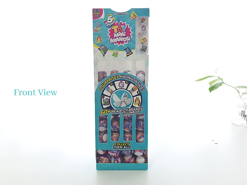 May include: A blue and white cardboard box with a display of small toy capsules. The box has the text "5 Mini Brands!" and "60+ Real Toy Brands to Unbox!" on it.