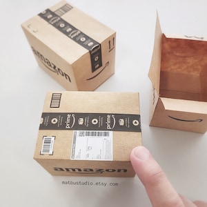 May include: Three brown cardboard boxes with the Amazon logo and Prime branding. The boxes are different sizes and one is open. The text on the box reads "amazon" and "matbustudio.etsy.com".