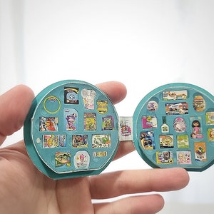 May include: A teal blue circular case with a hinged lid, open to reveal a variety of miniature items, including food, toys, and household products. The case is designed to resemble a miniature grocery store.