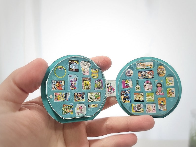 May include: A teal blue circular case with a hinged lid, open to reveal a variety of miniature items, including food, toys, and household products. The case is designed to resemble a miniature grocery store.