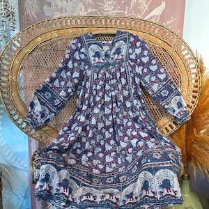 Könnte beinhalten: Ein blaues Kleid mit langen Ärmeln und einem Blumen- und Pfauenmuster. Das Kleid hat einen V-Ausschnitt und einen fließenden Rock. Der Stoff hat eine Mischung aus weißen, roten und blauen Farben. Das Kleid wird auf einem Korbstuhl präsentiert.