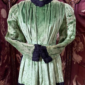 May include: A vintage green velvet dress with black trim and accents. The dress has a high collar, long sleeves, and a pleated skirt. Black trim is visible on the collar, cuffs, and hemline. The dress is displayed on a mannequin.