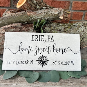 May include: A rectangular sign with the text "ERIE, PA home sweet home" in black script. Coordinates are printed below the text. The sign is light gray and rests on green leaves and logs.