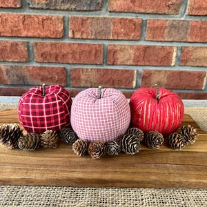 May include: Three fabric pumpkins in red plaid, pink gingham, and red with gold stripes, arranged on a wooden cutting board with pinecones. The pumpkins have wooden stems and are set against a brick wall.