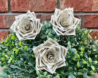 May include: Three white paper roses with musical notes printed on them are arranged in a green bush. The roses are made of paper and have a delicate, vintage look.