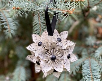 May include: A white paper ornament shaped like a flower with black buttons in the center. The ornament is hanging from a black ribbon on a green pine tree branch.