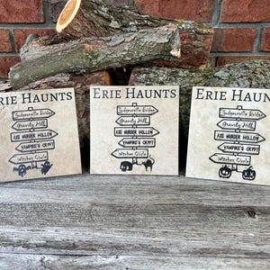 May include: Three square signs with the text "Erie Haunts" and directional arrows pointing to spooky locations like "Gravity Hill" and "Vampire's Crypt." The signs have a textured, beige background and black lettering. A log and brick wall are in the background.