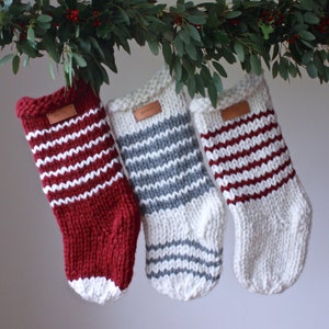 May include: Three knitted Christmas stockings with white and red stripes, white and grey stripes, and white and burgundy stripes. Each stocking has a leather tag with a name on it.