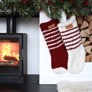 Peut inclure: Deux chaussettes de Noël en tricot rayées rouge et blanc avec une étiquette marron indiquant "Merry" accrochées à une cheminée blanche au-dessus d'un foyer.